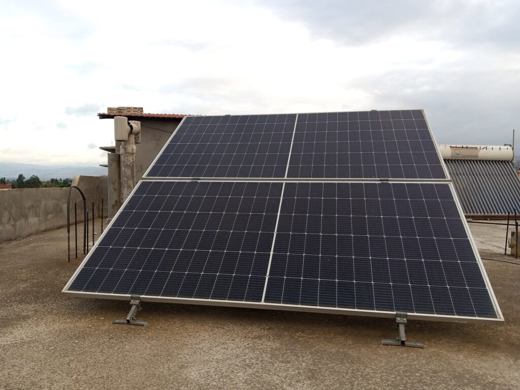 A close-up photograph of a residential off-grid photovoltaic array in rural Homs, Syria. The setup consists of 4